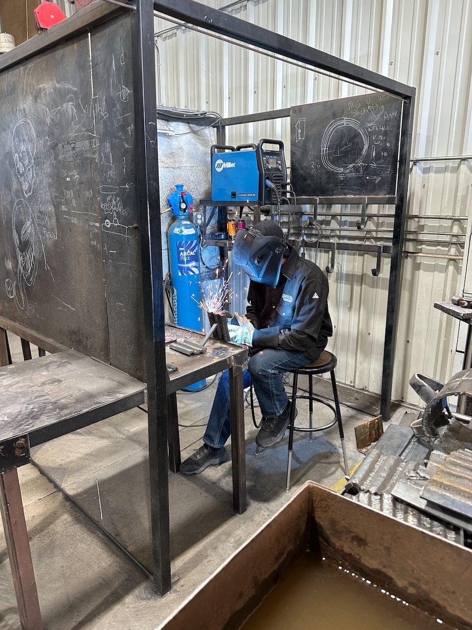 A learner wearing a welding helmet and gloves works at a metalworking station.